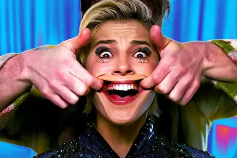 Manos estiran la boca de una mujer para crear una sonrisa forzada. Sus ojos están muy abiertos y maquillados.