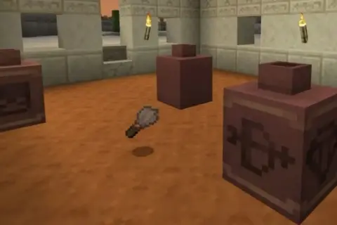 Un pincel de Minecraft flota sobre el suelo de arena, cerca de tres recipientes de arcilla con símbolos grabados.