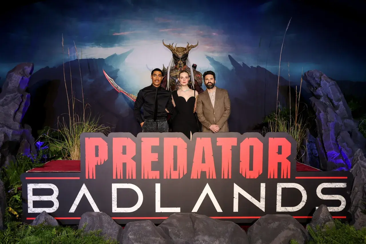 El reparto de Predator: Badlands posa frente a un telón de fondo dramático y un letrero.