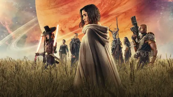 Un grupo de guerreros, incluyendo a una mujer con capa y espadas de luz, se alza en un campo de trigo bajo un cielo anaranjado y un planeta gigante. Son los rebeldes de Rebel Moon.