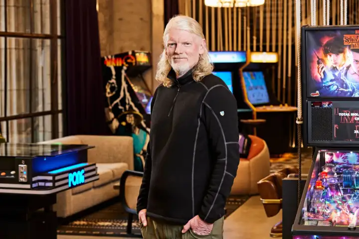 El CEO de Twitch, con su característico cabello rubio, posa sonriente rodeado de máquinas de arcade y un pinball de Stranger Things.